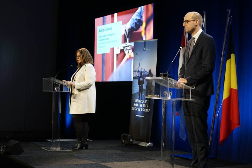 Staatssecretaris Aukje de Vries en de Belgische minister van Financiën Vincent van Peteghem presenteren samen de drugscijfers 2023 met op de achtergrond vlaggen en een presentatiescherm
